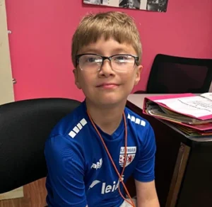 Blonde caucasian middle school aged boy with glasses wearing a soccer jersey, smiling at the camera