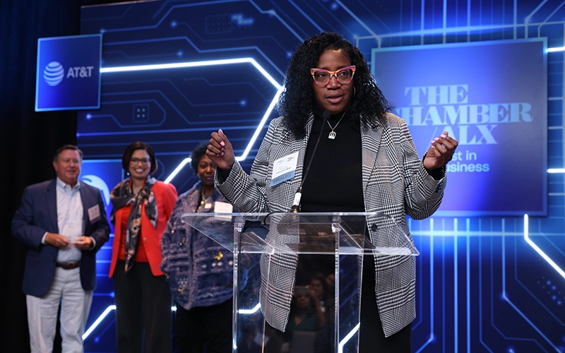 Professional Black woman wearing a black and white plaid blazer and stylish cat eye glasses gesturing at podium in front of a sign background saying The Chamber ALX: Best in Business