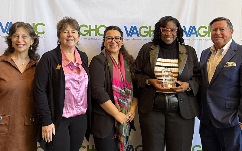 Four women and one man smiling in front of a VAGHC banner wall. One woman is holding an award trophy object.