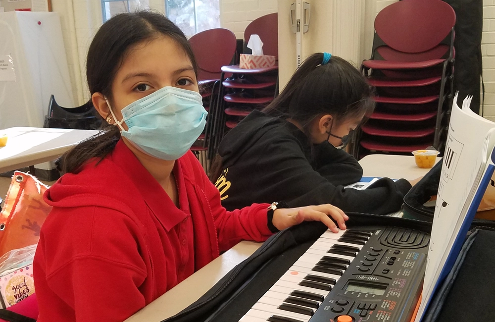 Young Latina girl practicing piano on a keyboard next to another Latina girl doing homework.