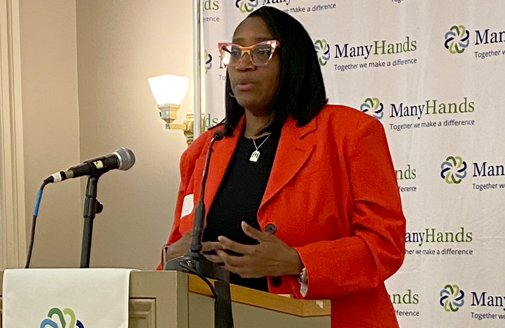 Professionally dressed Black woman speaking at a podium in front of a banner with the ManyHands logo repeated in the background.